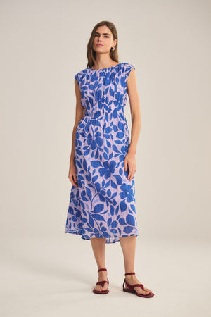 A woman wears the LEVAINE DRESS by Velvet by Graham & Spencer—a blue and white floral smocked waist dress with cap sleeves—paired with red flat sandals, standing against a plain light background.