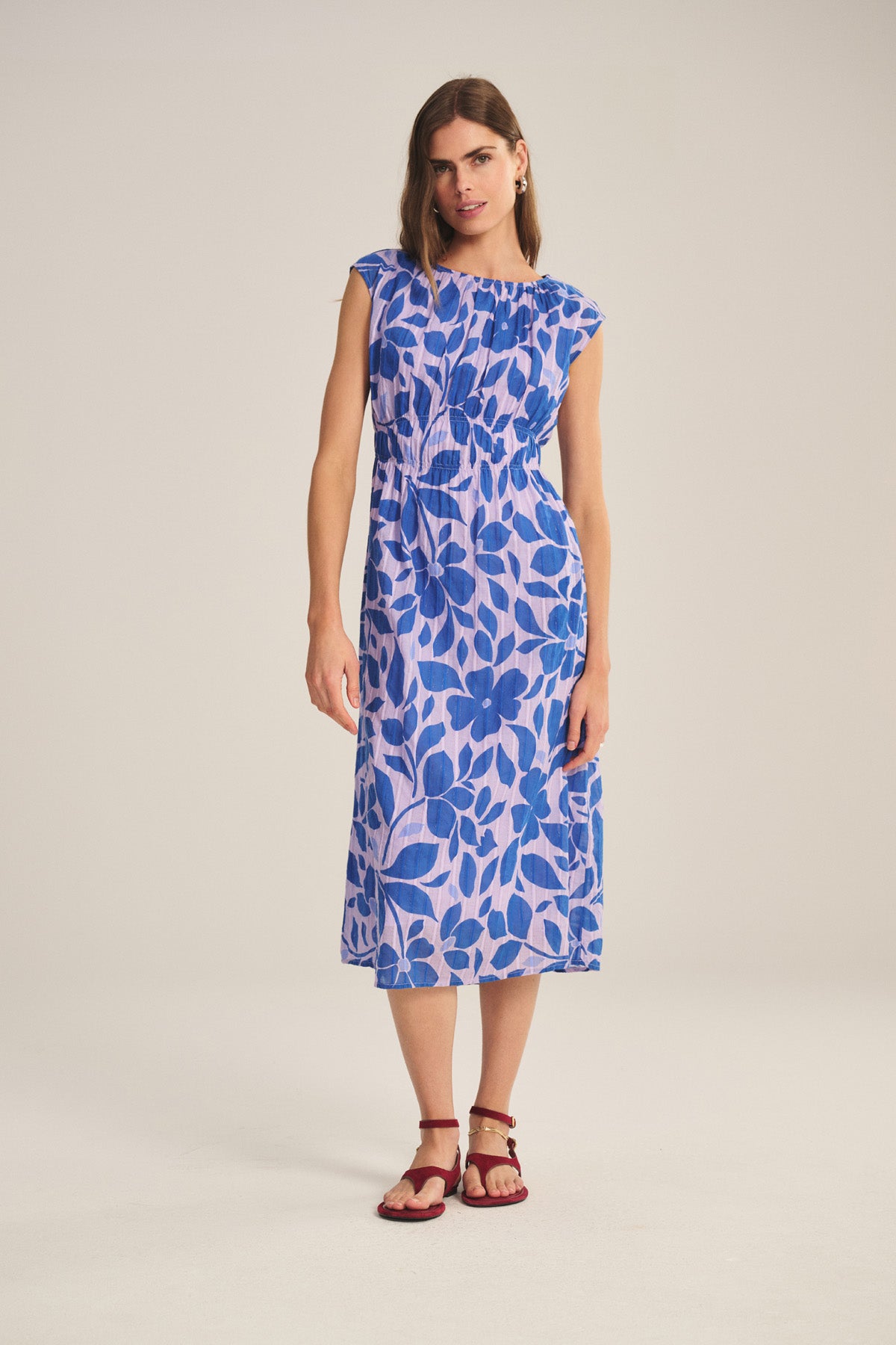 A woman wears the LEVAINE DRESS by Velvet by Graham & Spencer, a sleeveless blue and purple floral print cotton dress with a smocked waist, styled with burgundy sandals.