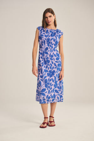A woman wears the LEVAINE DRESS by Velvet by Graham & Spencer, a sleeveless blue and purple floral print cotton dress with a smocked waist, styled with burgundy sandals.