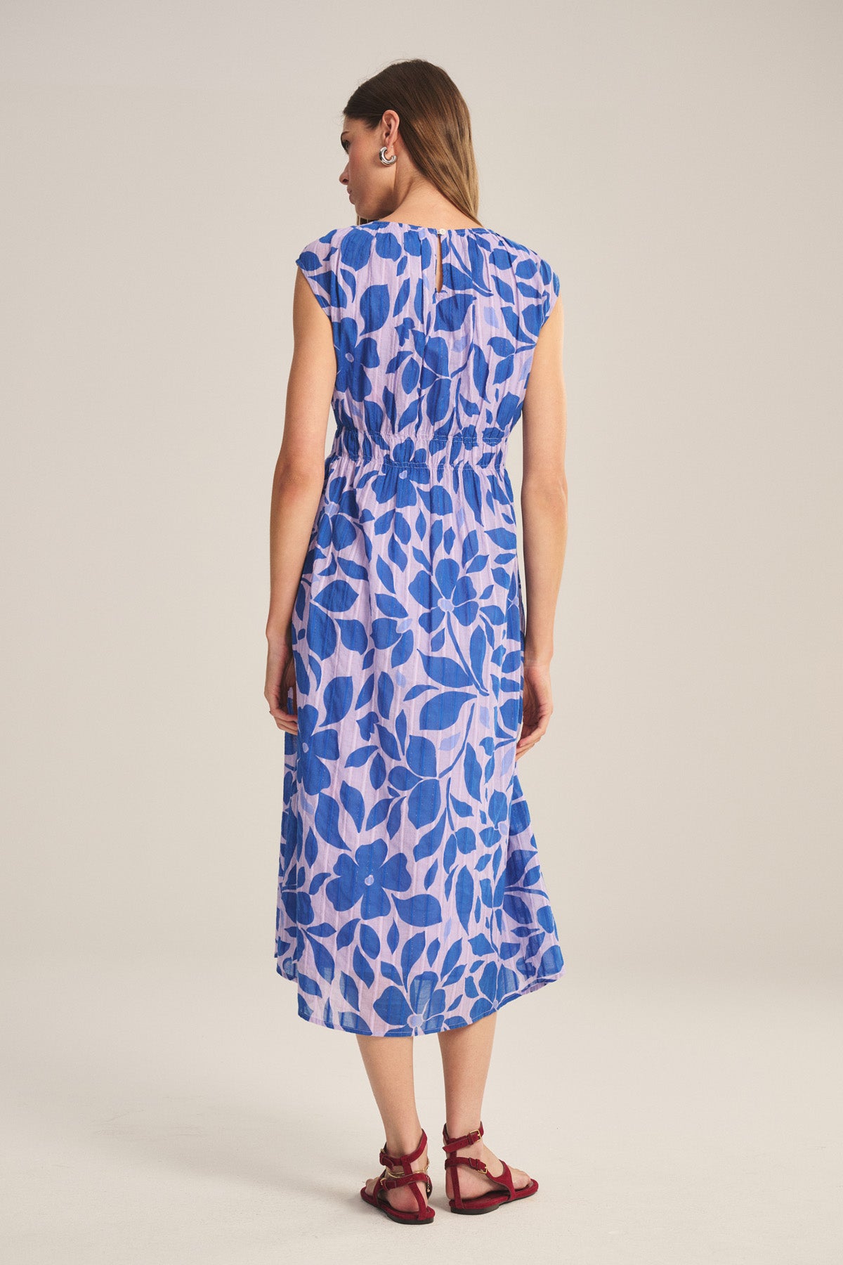A woman stands facing away, wearing the LEVAINE DRESS by Velvet by Graham & Spencer—a lightweight cotton dress with a blue floral pattern on a light background—paired with red sandals.-40808348549313