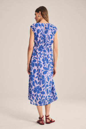 A woman stands facing away, wearing the LEVAINE DRESS by Velvet by Graham & Spencer—a lightweight cotton dress with a blue floral pattern on a light background—paired with red sandals.