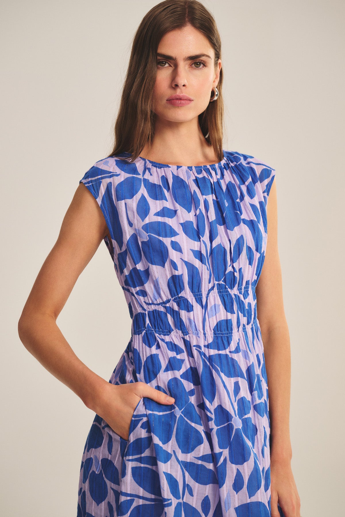 A woman with long brown hair wears the LEVAINE DRESS by Velvet by Graham & Spencer—a sleeveless, lightweight lilac cotton dress with a blue leaf pattern—standing against a neutral background, one hand in her pocket.-40808348516545