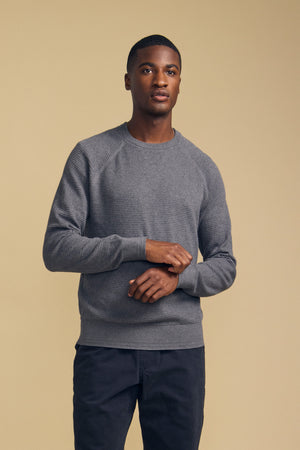 A man stands against a beige background wearing the AUGIE COTTON CASHMERE SWEATER by Velvet by Graham & Spencer—a gray crew-neck style—paired with dark pants, his hands loosely clasped in front of him.