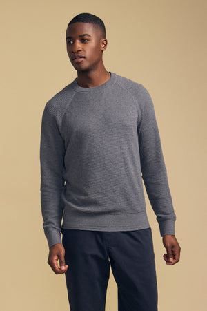 A man stands against a beige background wearing the Velvet by Graham & Spencer AUGIE COTTON CASHMERE SWEATER—a textured gray knit with a classic crew neckline—paired with dark pants, looking slightly to the side with a neutral expression.