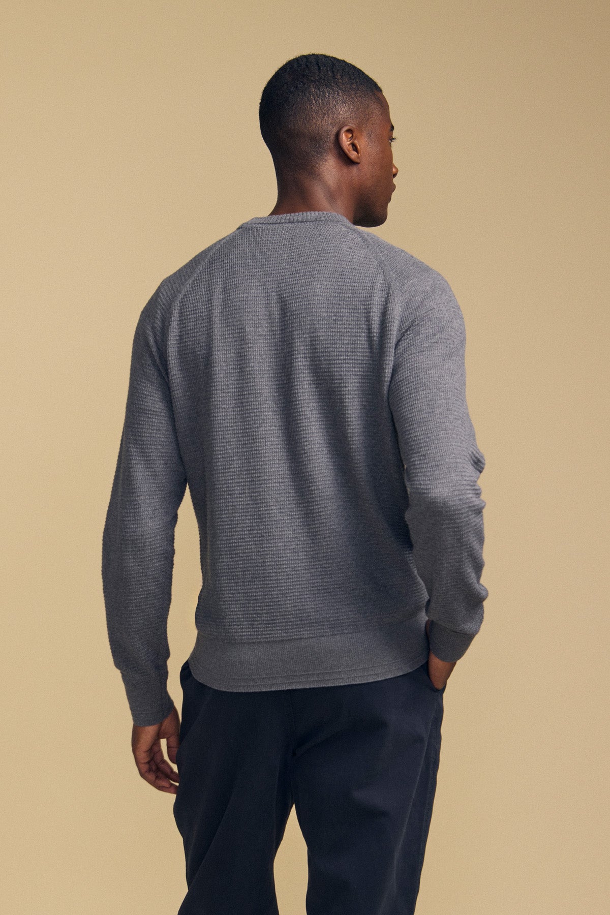 A man stands facing away, wearing the Velvet by Graham & Spencer AUGIE COTTON CASHMERE SWEATER—a textured gray crewneck—paired with dark pants against a plain beige background.