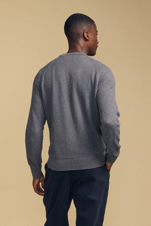 A man stands facing away, wearing the Velvet by Graham & Spencer AUGIE COTTON CASHMERE SWEATER—a textured gray crewneck—paired with dark pants against a plain beige background.