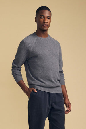 A man stands against a beige background wearing the Velvet by Graham & Spencer AUGIE Cotton Cashmere Sweater—a gray ribbed crewneck style—paired with dark trousers, one hand casually in his pocket.