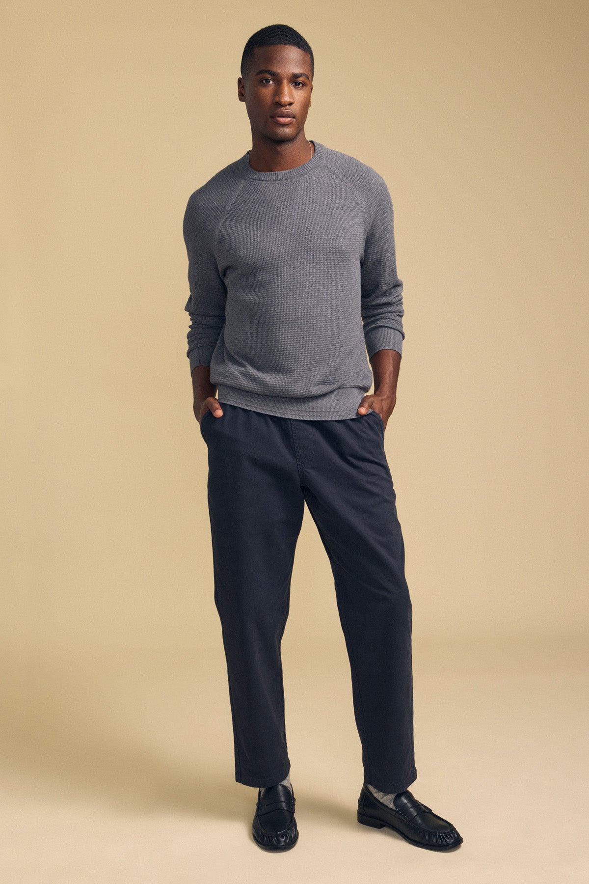 A man stands with his hands in his pockets, wearing the Velvet by Graham & Spencer AUGIE Cotton Cashmere Sweater in grey, featuring a classic crew neckline, paired with dark pants and black loafers against a beige background.