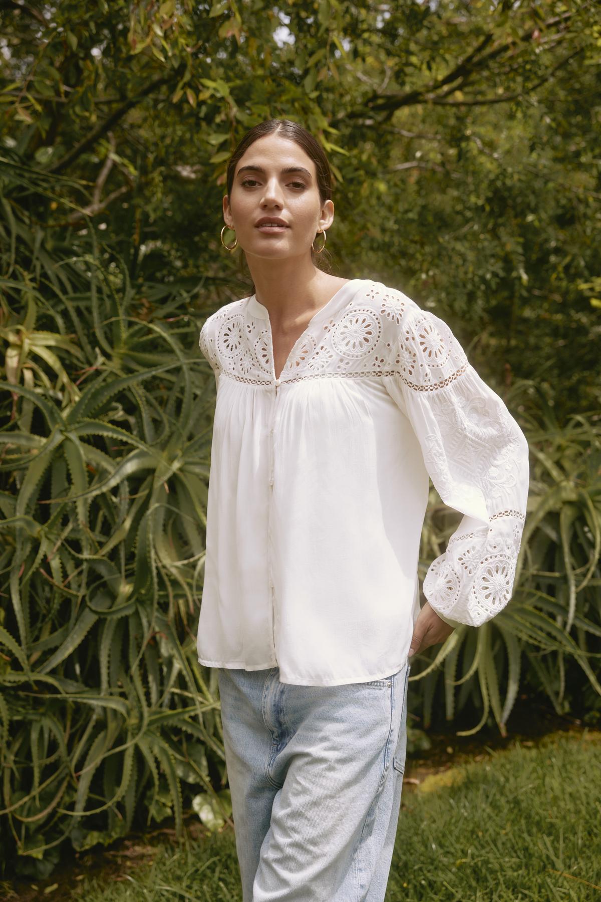   A person wearing the LIBBY TOP from Velvet by Graham & Spencer and jeans stands outdoors in front of greenery. 