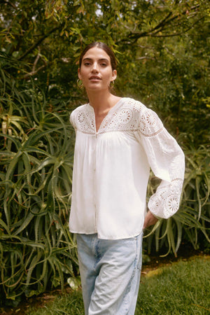 A person wearing the LIBBY TOP from Velvet by Graham & Spencer and jeans stands outdoors in front of greenery.