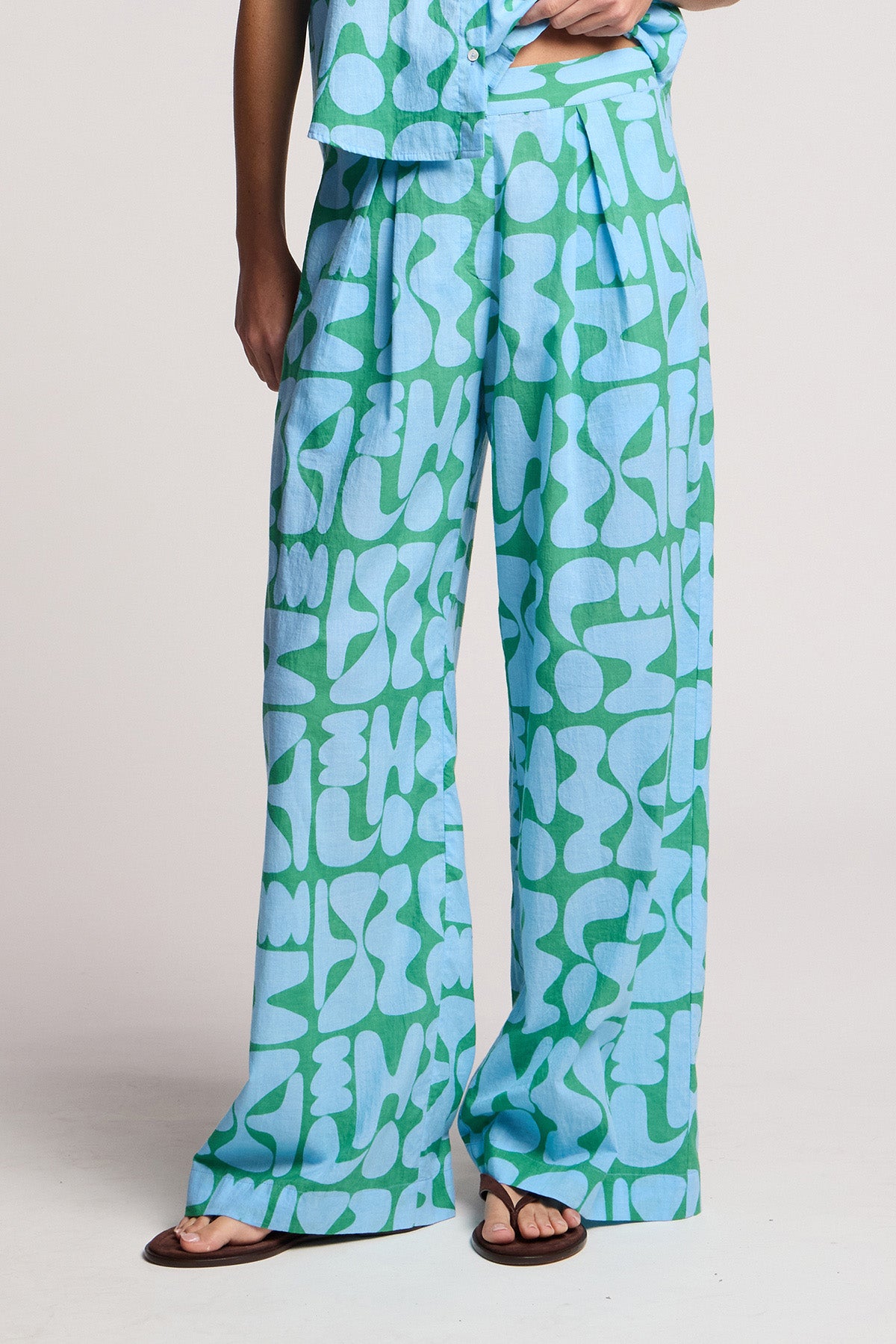 A person wearing the TRIXIE PANT by Velvet by Graham & Spencer—loose, high-waisted wide-leg pants in abstract green and light blue cotton cambric—paired with sandals and a matching shirt.