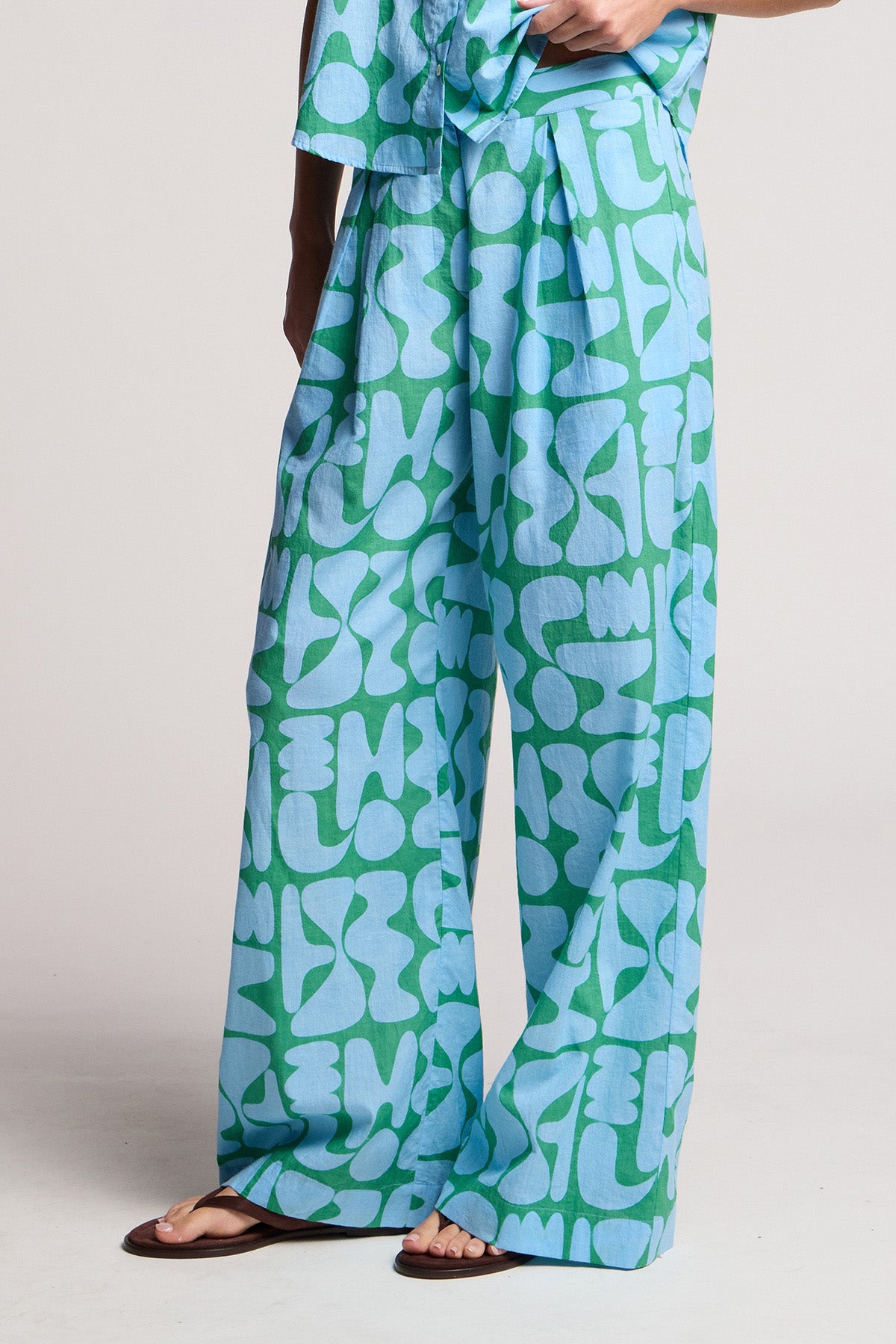 A person wearing the TRIXIE PANT by Velvet by Graham & Spencer—a wide-leg silhouette in blue and green abstract printed cotton cambric—stands against a plain light background, paired with brown sandals.