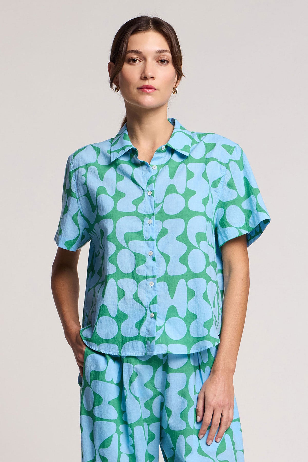 A woman wears the MARINE SHIRT by Velvet by Graham & Spencer— a short-sleeve cotton cambric resort shirt with matching blue and green abstract patterned pants, standing against a plain background.-41645082050753