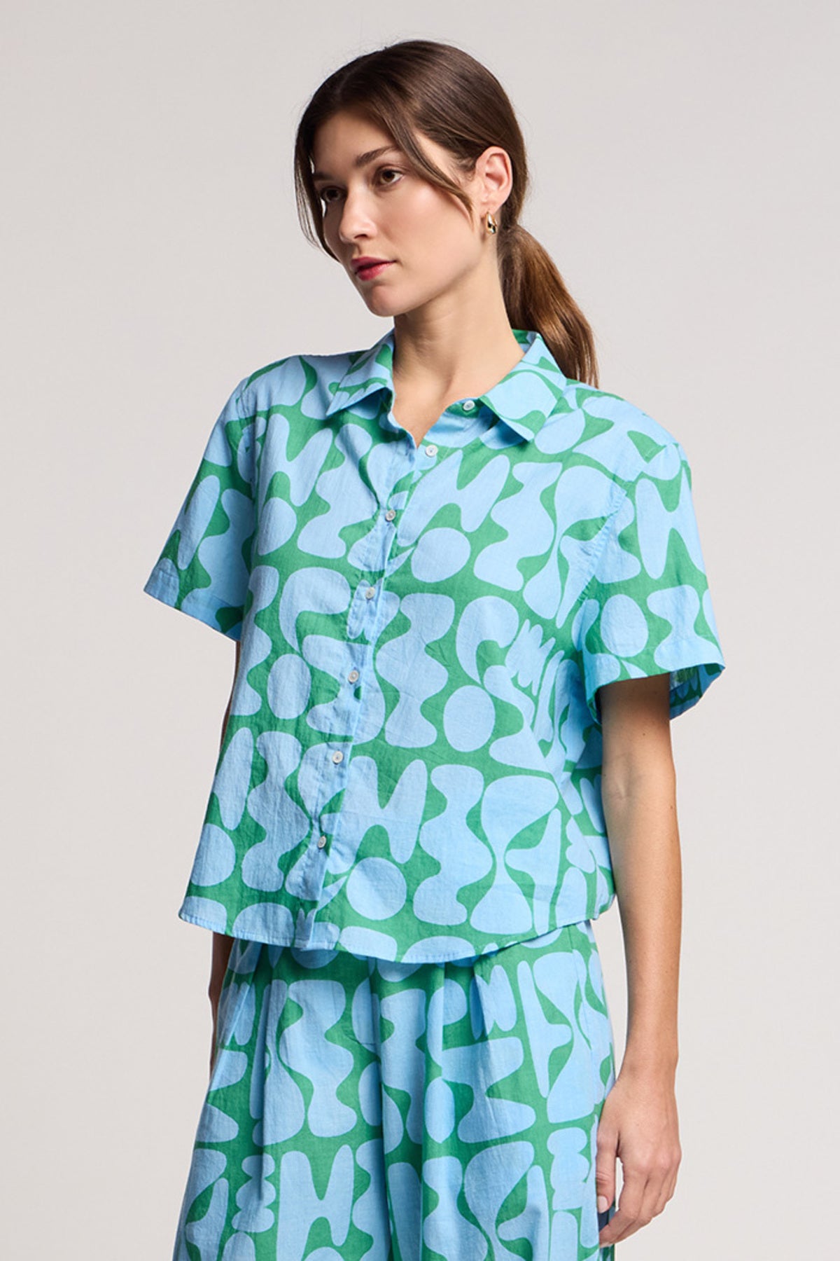 A person wears the Velvet by Graham & Spencer MARINE SHIRT and matching shorts in a light blue and green pattern, crafted from soft cotton cambric, standing against a plain background.-41645083787457