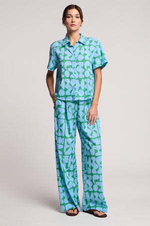A person wears the MARINE SHIRT by Velvet by Graham & Spencer, paired with matching wide-leg pants featuring a blue and green abstract pattern; they complete the look with sandals and a neutral expression.
