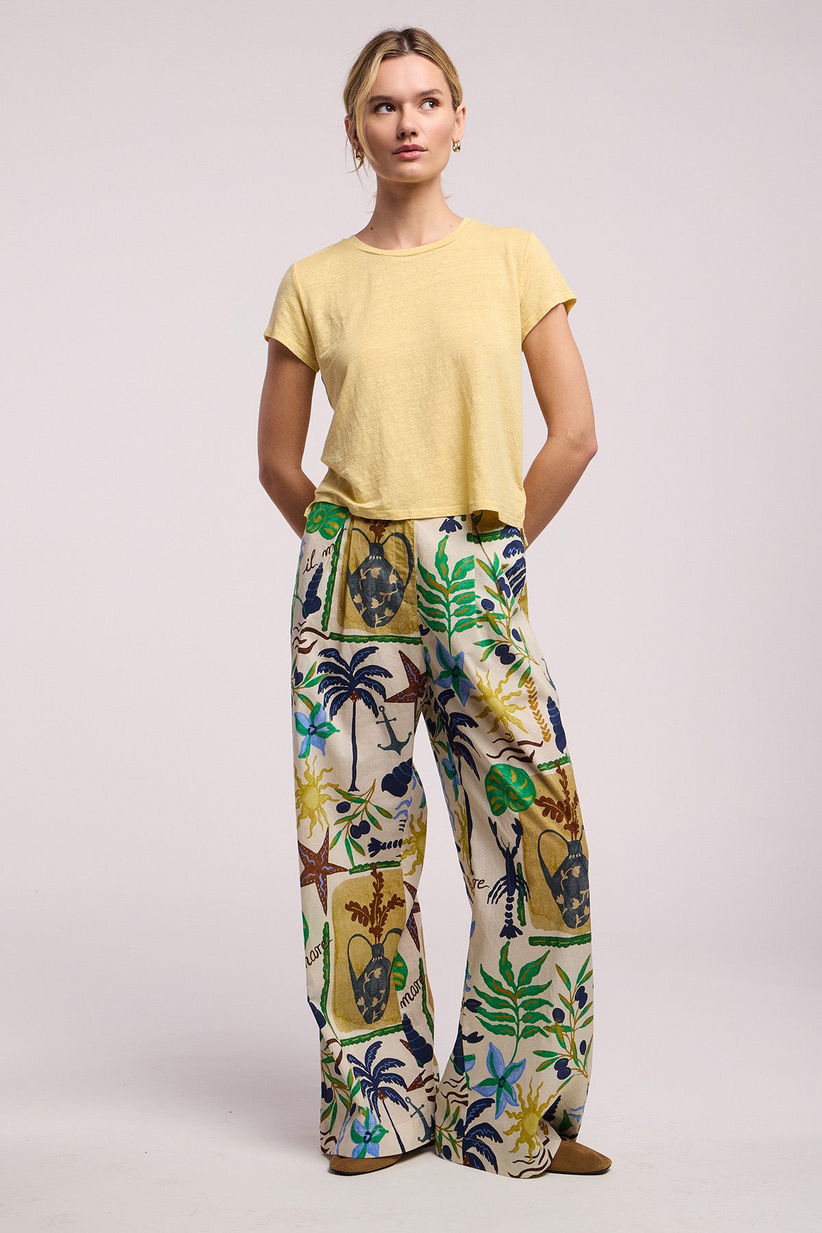 A woman stands against a plain background wearing Velvet by Graham & Spencer's TRIXIE PANT, a cotton cambric wide-leg statement pant with a vibrant tropical leaf and mask print, paired with a short-sleeve yellow top.