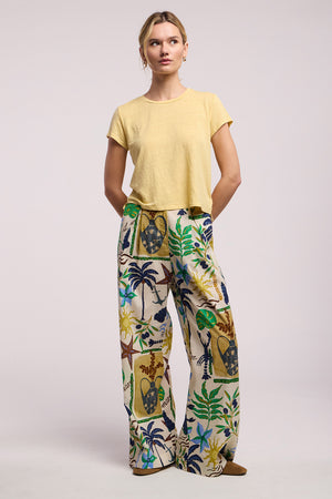 A woman stands against a plain background wearing Velvet by Graham & Spencer's TRIXIE PANT, a cotton cambric wide-leg statement pant with a vibrant tropical leaf and mask print, paired with a short-sleeve yellow top.