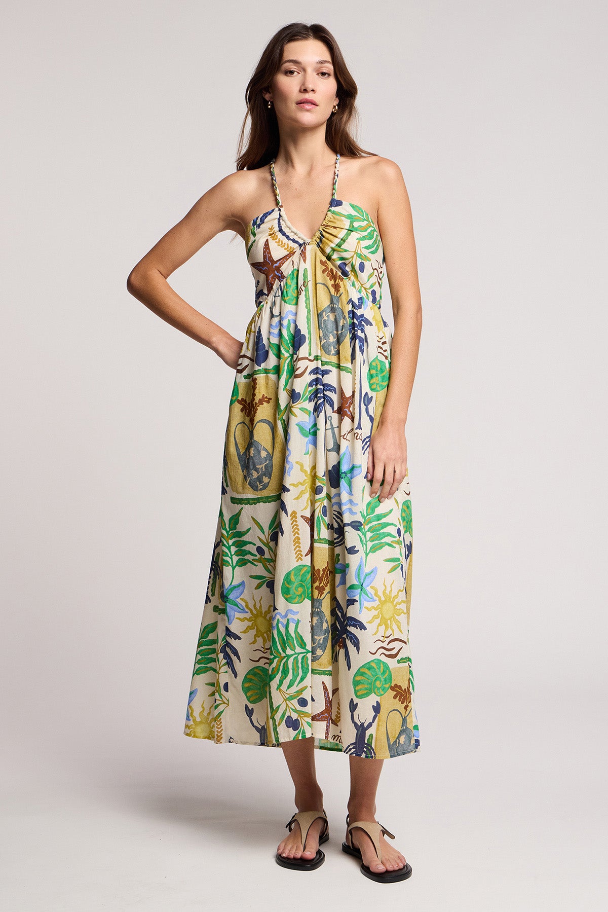 A woman stands against a plain background wearing the LEYLA DRESS by Velvet by Graham & Spencer—a sleeveless cotton voile maxi with braided halter straps and a vibrant botanical and animal print, styled with black sandals.