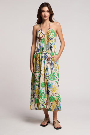 A woman wears the Velvet by Graham & Spencer LEYLA DRESS, a sleeveless printed cotton voile maxi with braided halter straps and a colorful botanical and animal print, paired with black sandals against a plain background.
