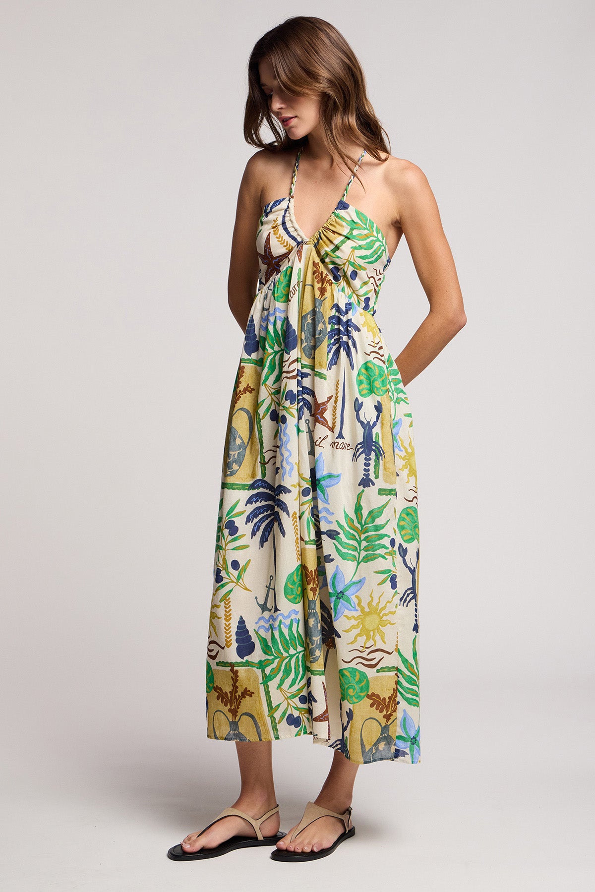 A woman wears the LEYLA DRESS by Velvet by Graham & Spencer—a colorful, sleeveless tropical-print maxi with braided halter straps—paired with black sandals, standing against a plain light background.-41527368712385