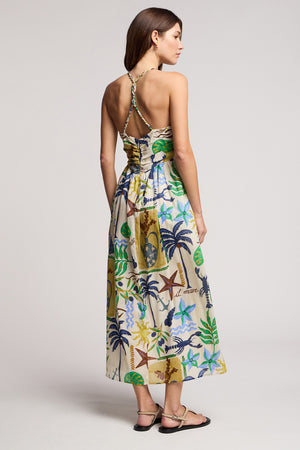 A woman stands facing away in the LEYLA DRESS by Velvet by Graham & Spencer—a sleeveless, printed cotton voile maxi with braided halter straps—paired with flat sandals.