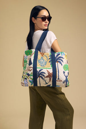 A woman with long dark hair, wearing sunglasses, a white t-shirt, and olive pants, carries the Velvet by Graham & Spencer LIDO PRINTED TOTE—a large tote bag featuring vibrant tropical prints—over her shoulder against a neutral beige background.