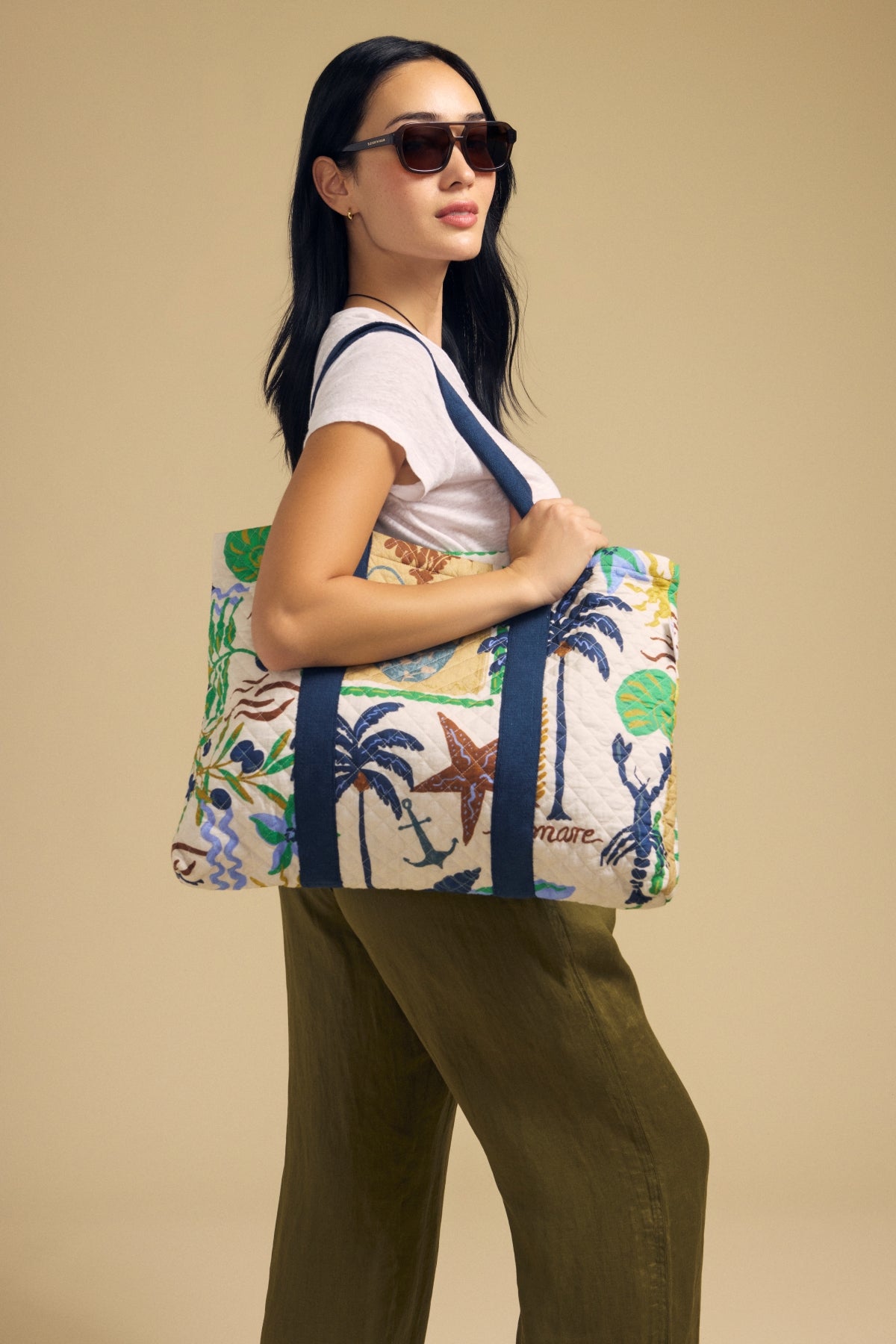 A woman wearing sunglasses and a white t-shirt holds the Velvet by Graham & Spencer LIDO PRINTED TOTE, featuring a bold tropical pattern, over her shoulder as she stands against a neutral background.