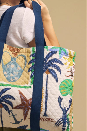 A woman wearing sunglasses holds the LIDO PRINTED TOTE by Velvet by Graham & Spencer—a large quilted bag featuring nautical and botanical motifs like palm trees, shells, and starfish—against a plain beige background.