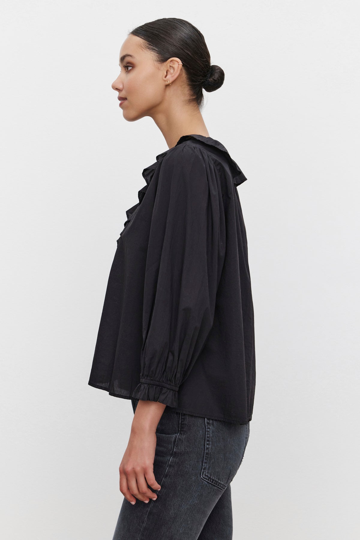   Against a plain white background, a person in profile wears the CINTHIA BLOUSE by Velvet by Graham & Spencer, featuring a black cotton poplin fabric with a ruffled collar and puffed sleeves, paired with dark jeans. 