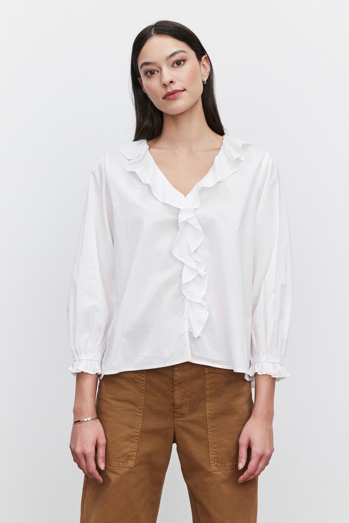   A person with long dark hair wears the CINTHIA BLOUSE, a white lightweight cotton poplin blouse with a ruffle placket from Velvet by Graham & Spencer, paired with brown pants, standing against a plain white background. 