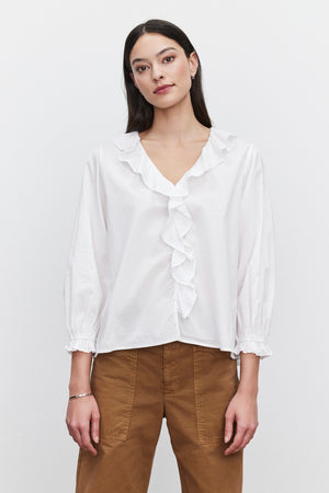 A person with long dark hair wears the CINTHIA BLOUSE, a white lightweight cotton poplin blouse with a ruffle placket from Velvet by Graham & Spencer, paired with brown pants, standing against a plain white background.