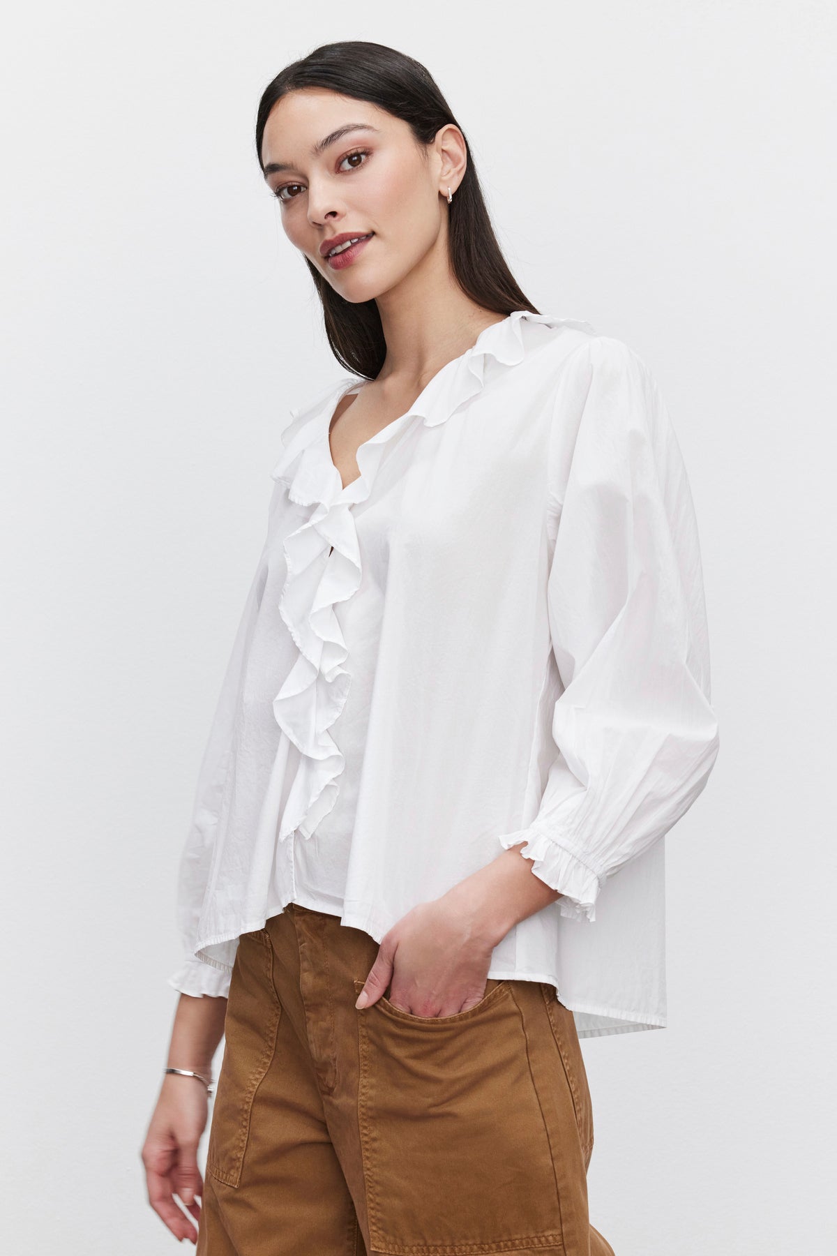   A person wearing the CINTHIA BLOUSE from Velvet by Graham & Spencer, made of lightweight cotton poplin with a ruffle placket and three-quarter sleeves, paired with brown pants, stands against a plain background. 