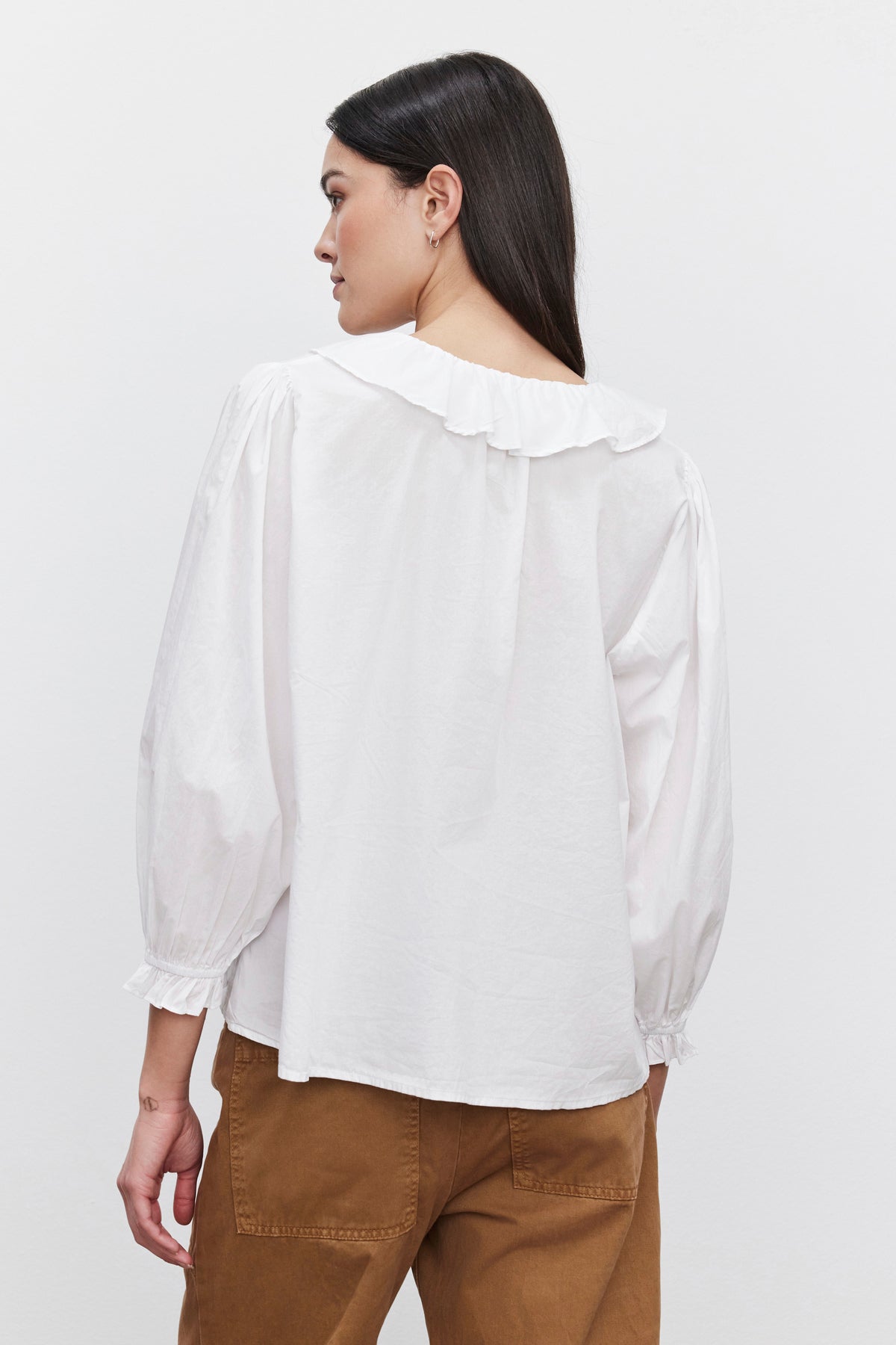   A person with long dark hair is standing, facing away, wearing the CINTHIA BLOUSE by Velvet by Graham & Spencer, which features a white long-sleeve design with a ruffled collar, paired with brown pants. The lightweight cotton poplin fabric of the blouse adds an understated elegance against the plain white background. 