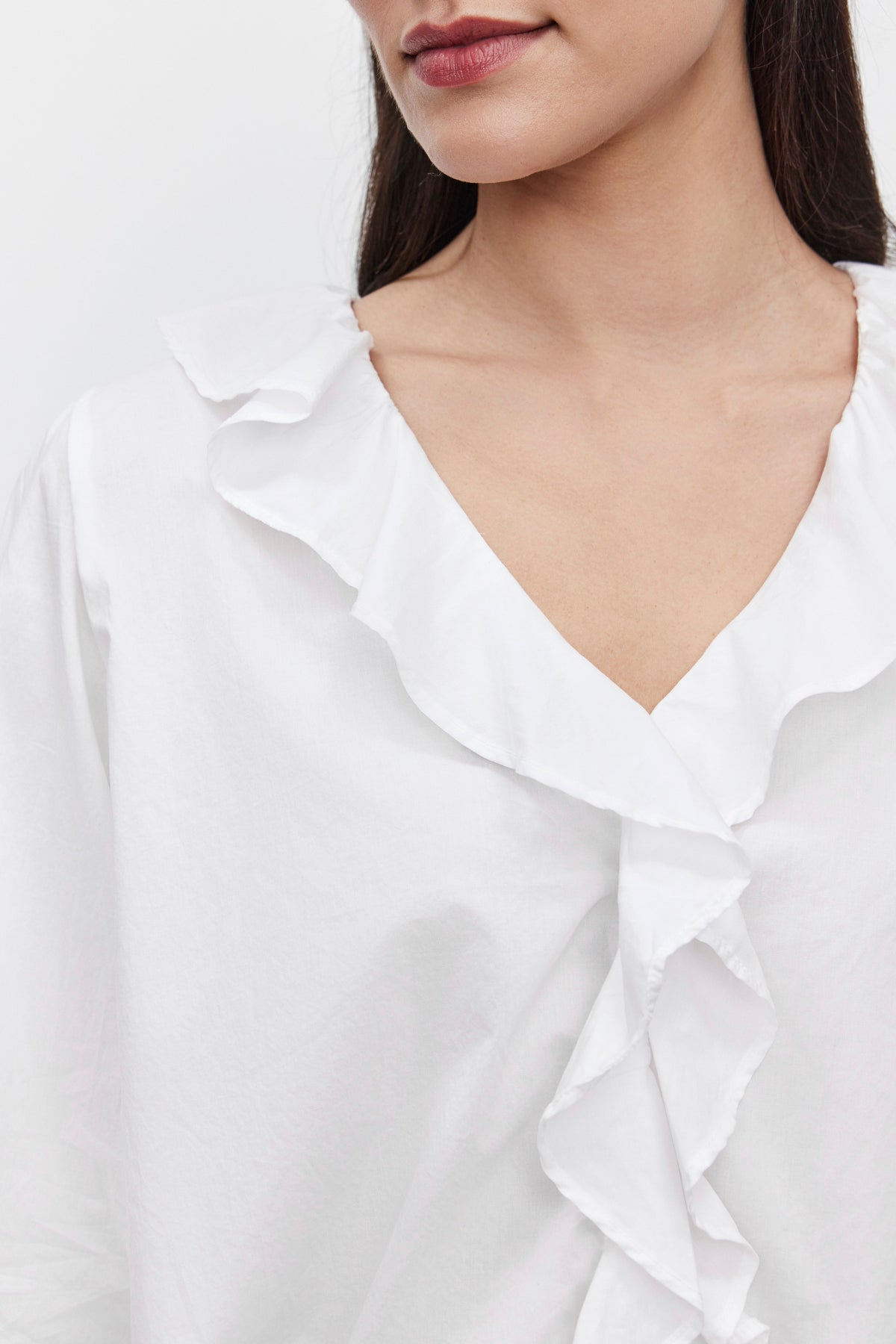   Close-up of a person wearing the CINTHIA BLOUSE from Velvet by Graham & Spencer, crafted from lightweight cotton poplin and featuring a V-neckline with an elegant ruffle placket. The individual has dark hair and is shown from the nose down, set against a plain background. 