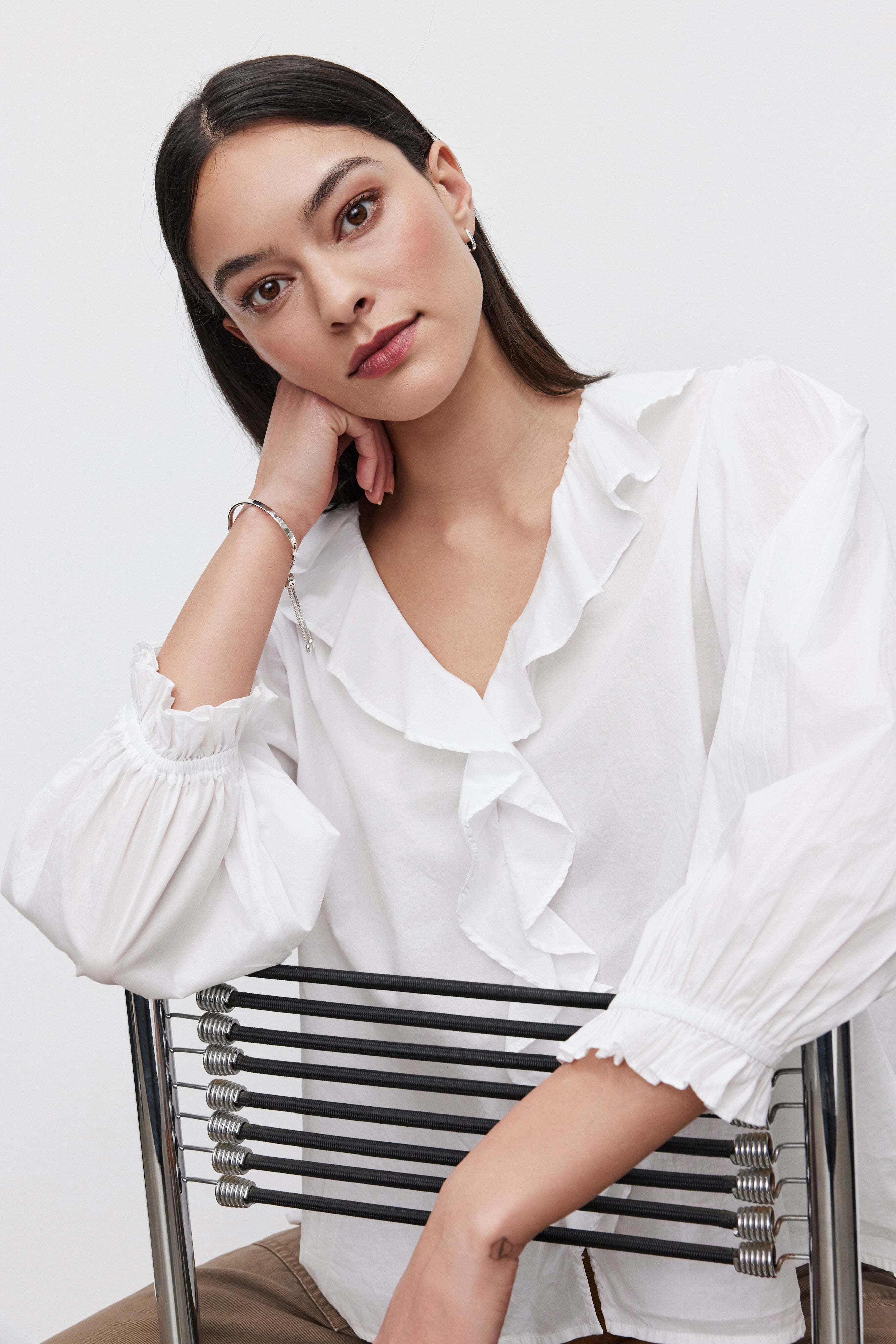   A woman with dark hair, dressed in a Velvet by Graham & Spencer CINTHIA BLOUSE featuring a lightweight cotton poplin fabric and ruffle placket, rests her head on her hand while seated on a metallic chair. 