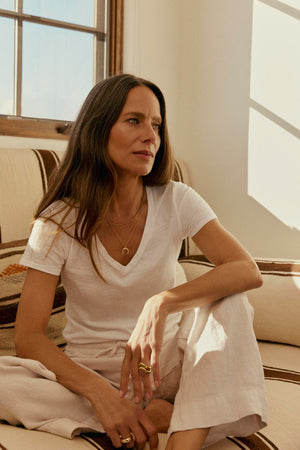 Woman in a Gwyneth Heavy Linen Pant by Velvet by Graham & Spencer, white outfit sitting and gazing thoughtfully by the window, with sunlight casting shadows in the room.