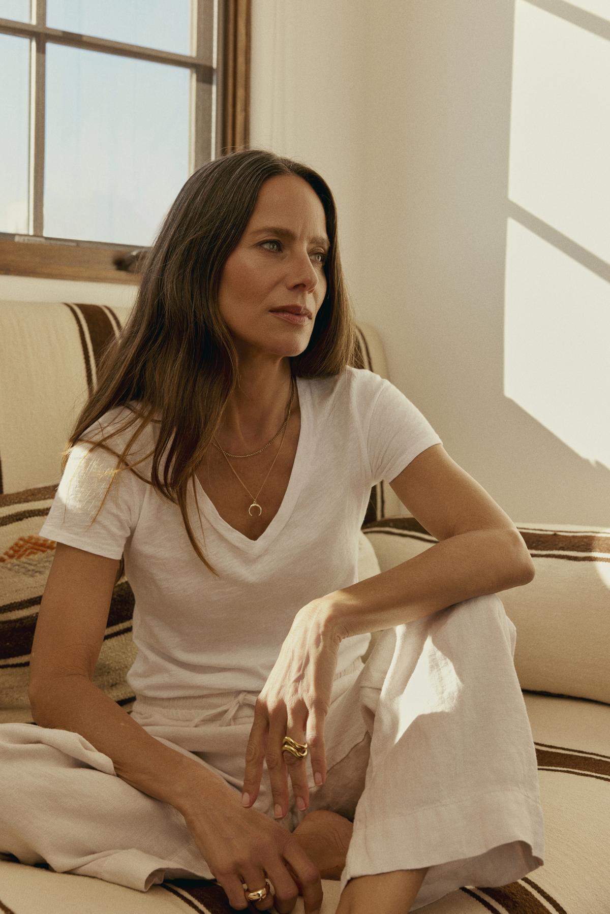   A woman in a Velvet by Graham & Spencer LILITH COTTON SLUB V-NECK TEE and pants sits cross-legged on a couch, looking thoughtful, with sunlight casting shadows in the room. 