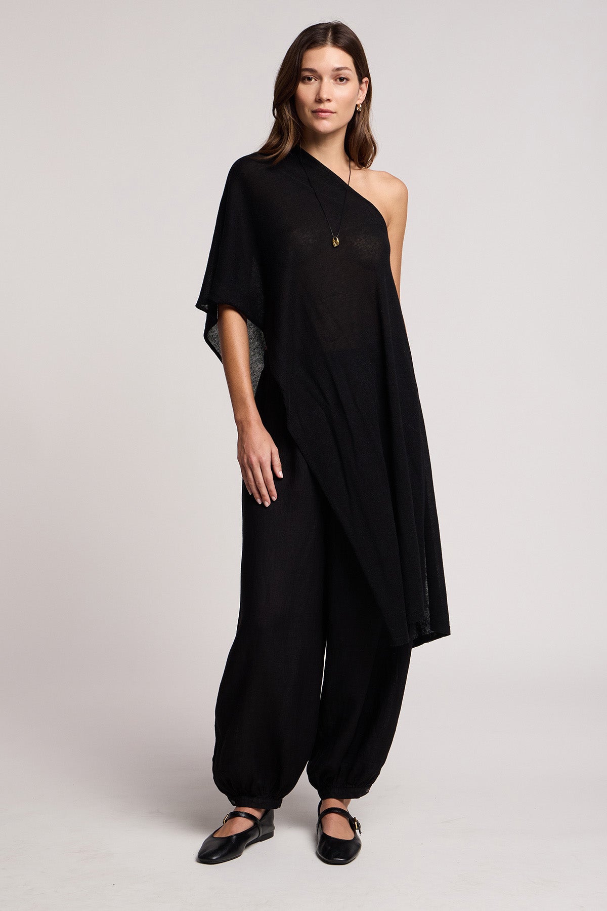 A woman stands facing forward against a plain light background, wearing the FLOREN SWEATER by Velvet by Graham & Spencer—a black one-shoulder asymmetrical top—paired with loose black pants, black flat shoes, and a necklace.