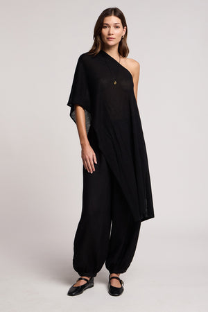 A woman stands facing forward against a plain light background, wearing the FLOREN SWEATER by Velvet by Graham & Spencer—a black one-shoulder asymmetrical top—paired with loose black pants, black flat shoes, and a necklace.