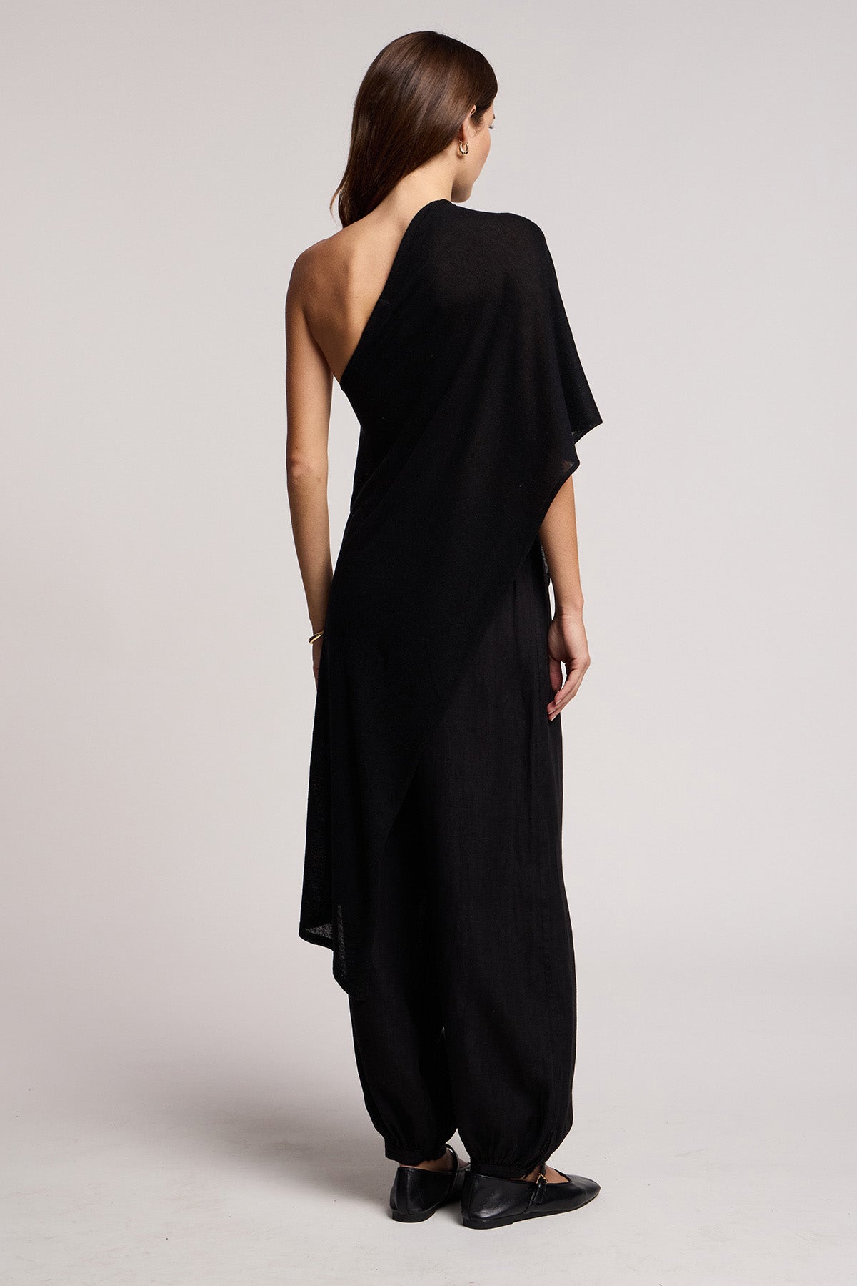 A woman faces away, wearing the FLOREN SWEATER by Velvet by Graham & Spencer in black with one-shoulder, draped detailing and paired with black shoes, set against a plain background.