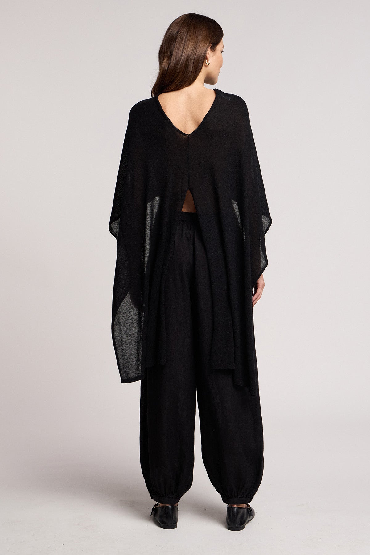 A woman stands with her back to the camera, wearing the Velvet by Graham & Spencer FLOREN SWEATER—a loose black top with a V-shaped back and wide sleeves—paired with black, loose-fitting pants and black shoes.-41371932688577