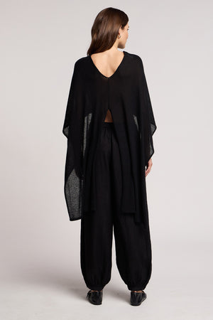 A woman stands with her back to the camera, wearing the Velvet by Graham & Spencer FLOREN SWEATER—a loose black top with a V-shaped back and wide sleeves—paired with black, loose-fitting pants and black shoes.
