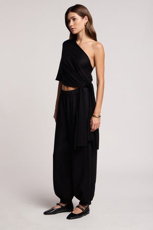 A woman stands in a studio, wearing the FLOREN SWEATER by Velvet by Graham & Spencer as a black one-shoulder top, paired with loose black pants and flat shoes, facing slightly to the left.