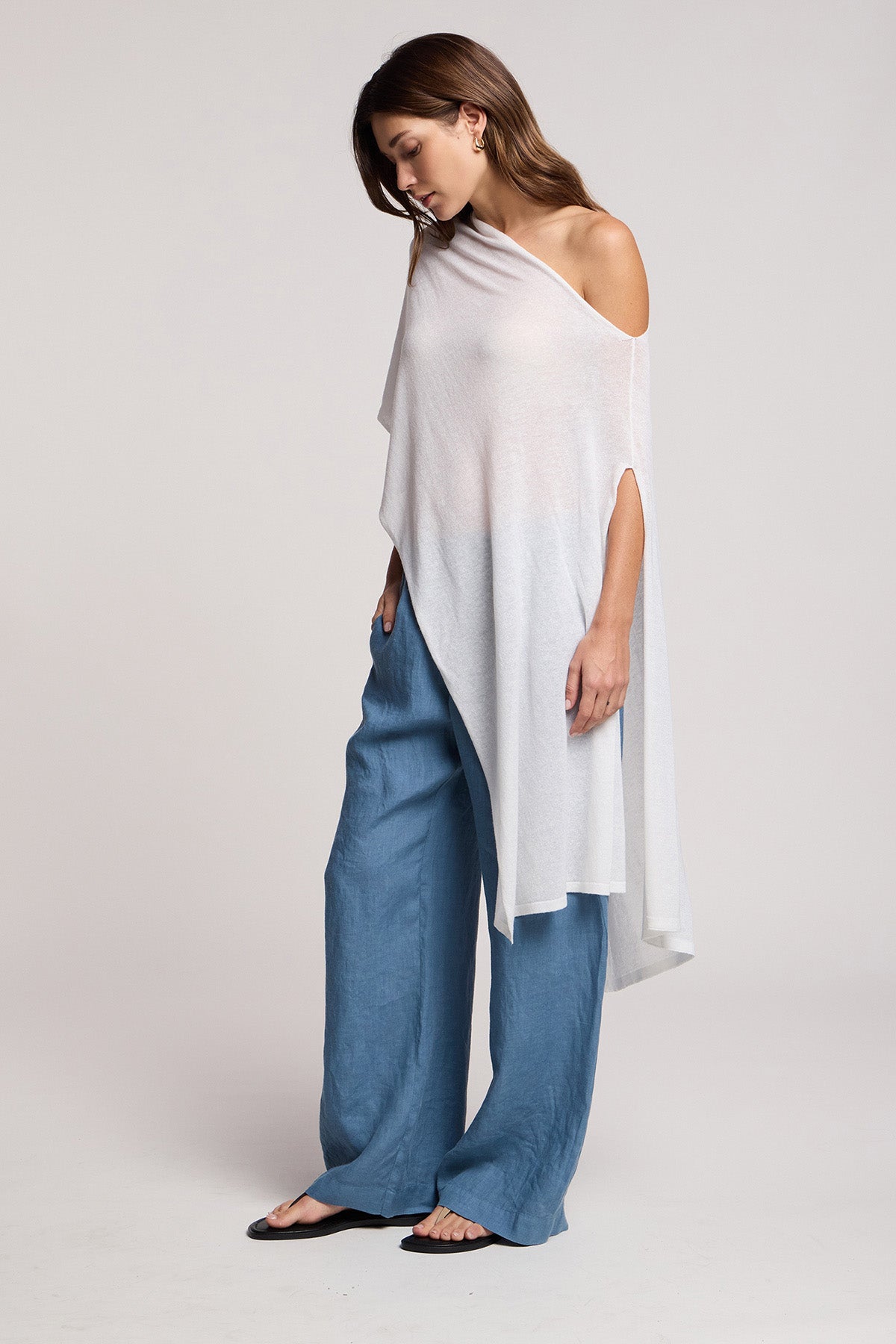 A woman wearing the FLOREN SWEATER by Velvet by Graham & Spencer, an asymmetrical white one-shoulder top, pairs it with loose blue pants and flat black sandals, standing against a plain light background and looking down.-41371932262593