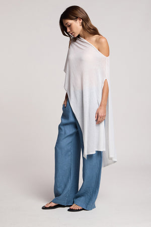 A woman wearing the FLOREN SWEATER by Velvet by Graham & Spencer, an asymmetrical white one-shoulder top, pairs it with loose blue pants and flat black sandals, standing against a plain light background and looking down.