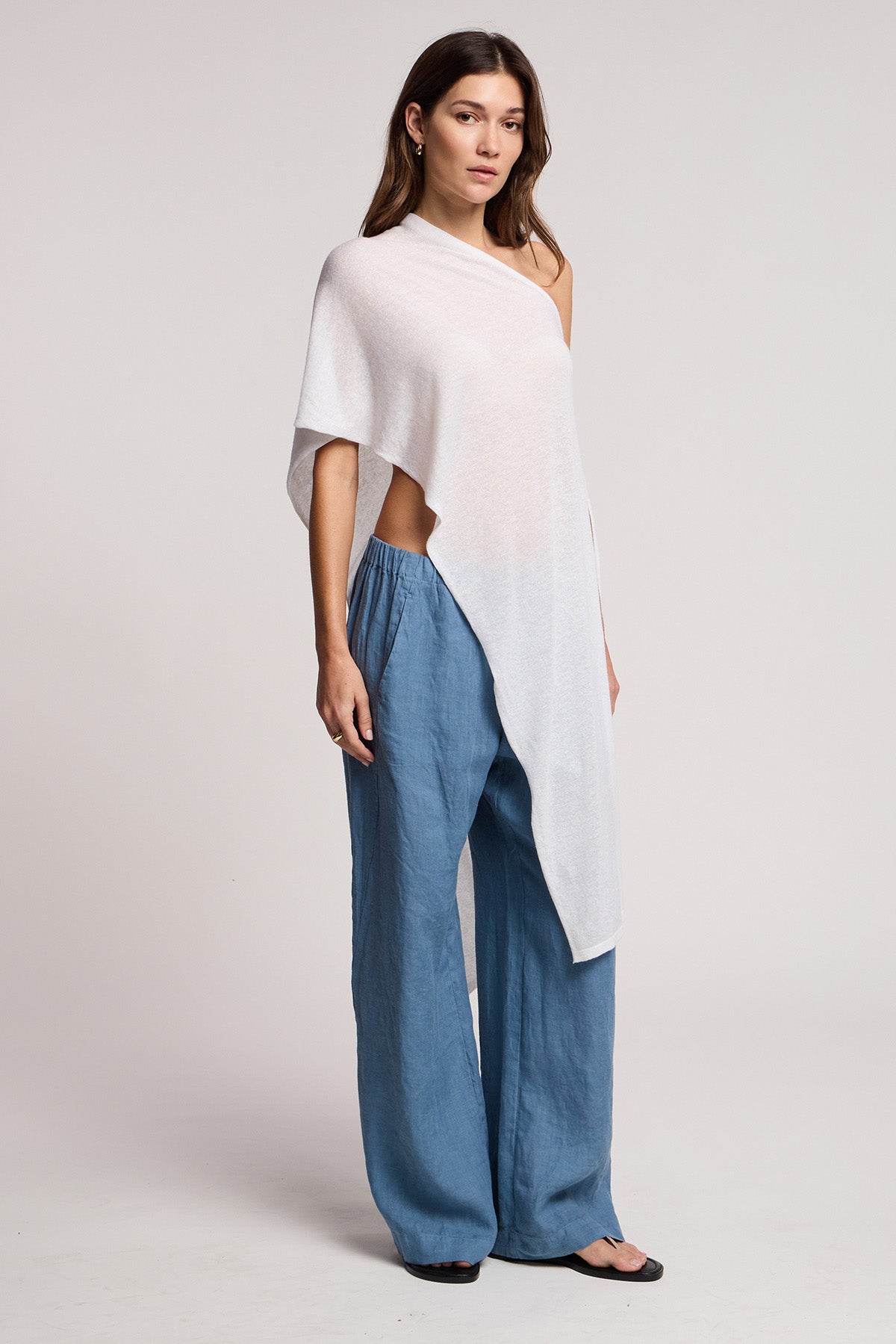 A woman stands in a studio wearing the FLOREN SWEATER by Velvet by Graham & Spencer, paired with loose blue pants and black sandals. She faces forward with a neutral expression.