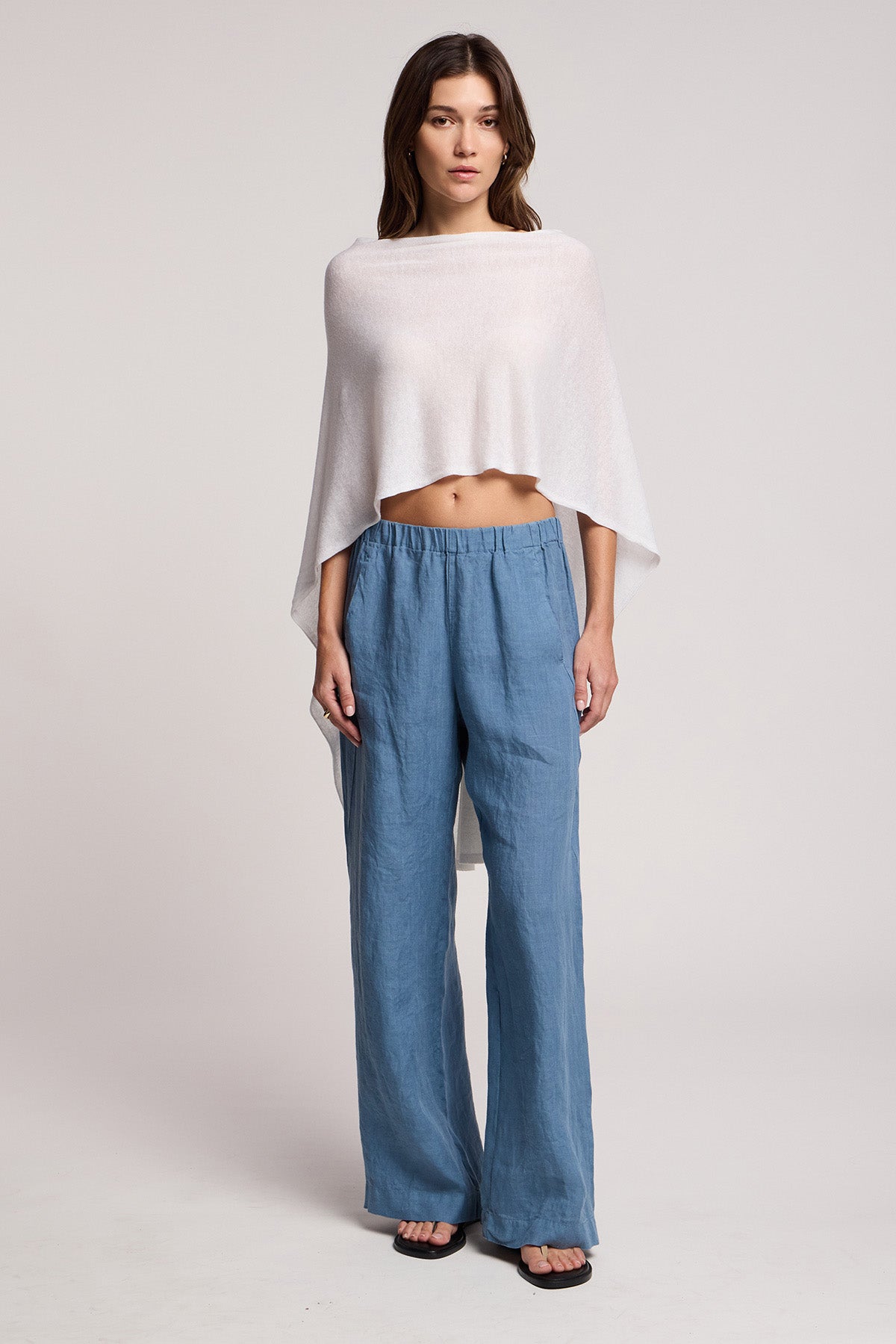 A woman faces forward against a plain background, wearing the FLOREN SWEATER by Velvet by Graham & Spencer—a light, loose-fitting white top—paired with blue wide-leg pants and sandals.-41371931934913