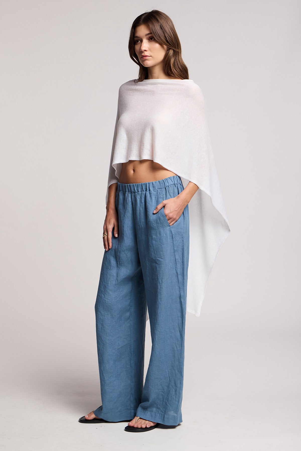 Woman standing in a studio wearing the FLOREN SWEATER by Velvet by Graham & Spencer, styled as an off-shoulder white poncho top, paired with loose blue pants and black sandals, one hand in her pocket.-41371932000449