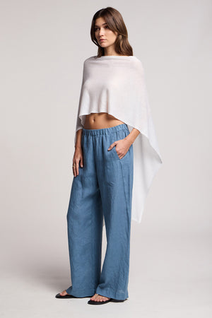 Woman standing in a studio wearing the FLOREN SWEATER by Velvet by Graham & Spencer, styled as an off-shoulder white poncho top, paired with loose blue pants and black sandals, one hand in her pocket.