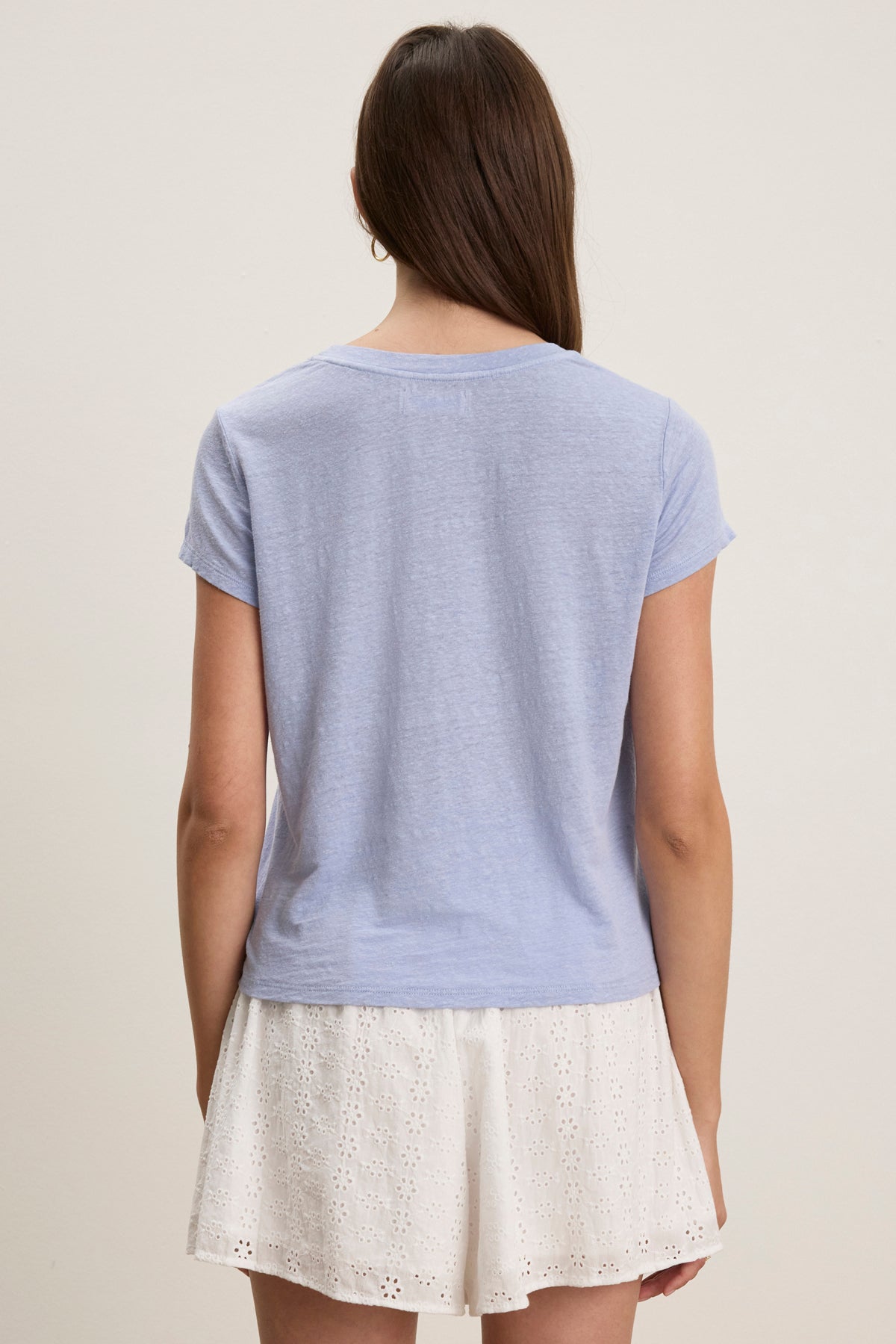 A person with long brown hair is seen from the back wearing the Velvet by Graham & Spencer CASEY LINEN KNIT TEE in light blue, paired with white eyelet shorts, set against a plain background.-39123274825921